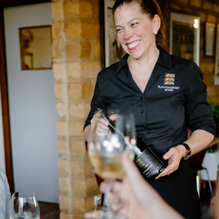 Woman in a black shirt with a logo, holding a wine bottle and glass in an indoor setting.