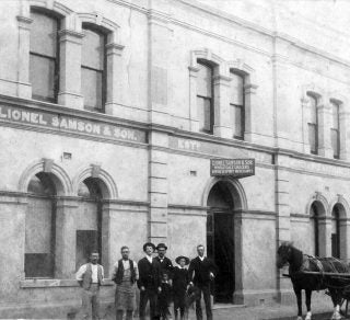 Historical black and white photo of a group of men standing outside a building with 'Lionel Sambon & Co' sign.
