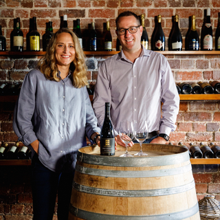 Two people standing behind a wooden barrel with wine bottles in the background.