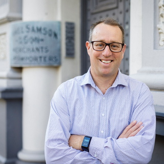 Man wearing glasses and a light blue shirt standing in front of a building with a sign.