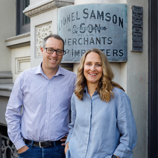 Tom and Jo Wisdom standing in front of a building with a sign that reads 'Lionel Samson & Son'.