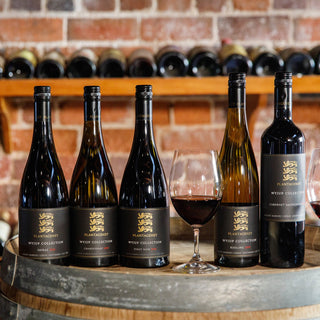Wine bottles on a wooden barrel with wine glasses, against a brick wall backdrop.